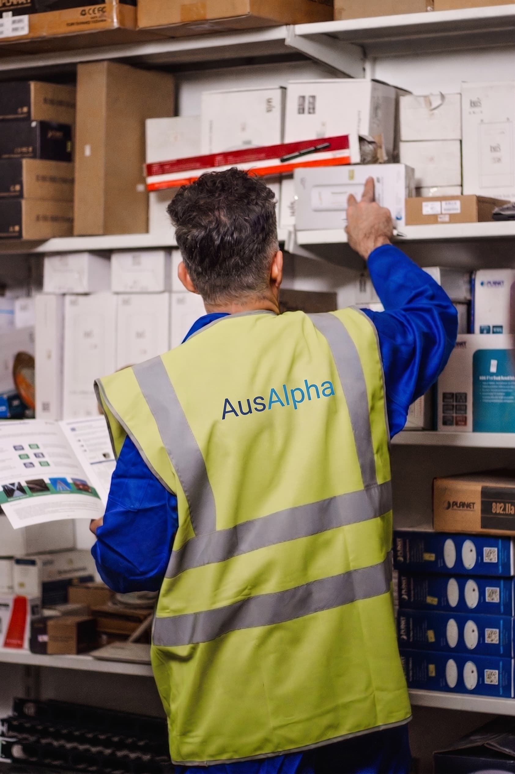 An AusAlpha team member in a branded hi-vis vest checking boxed stock against a printed list in the storeroom